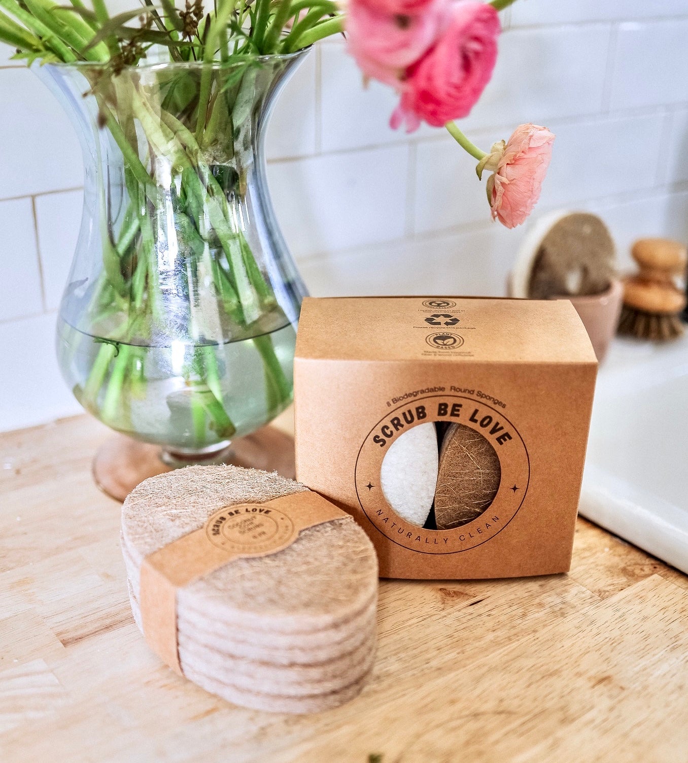 image of scrub be love toughest scrubbing coconut coir scrubbies and a box of the world's best oval dish sponges next to a vase of various shades of pink ranunculus flowers all sitting on a butcher block counter next to a sink. Bamboo scrub brush, mauve diatomite sponge holder holding a round Scrub Be Love sponge with heart cut out. Scrub Daddy Alternative