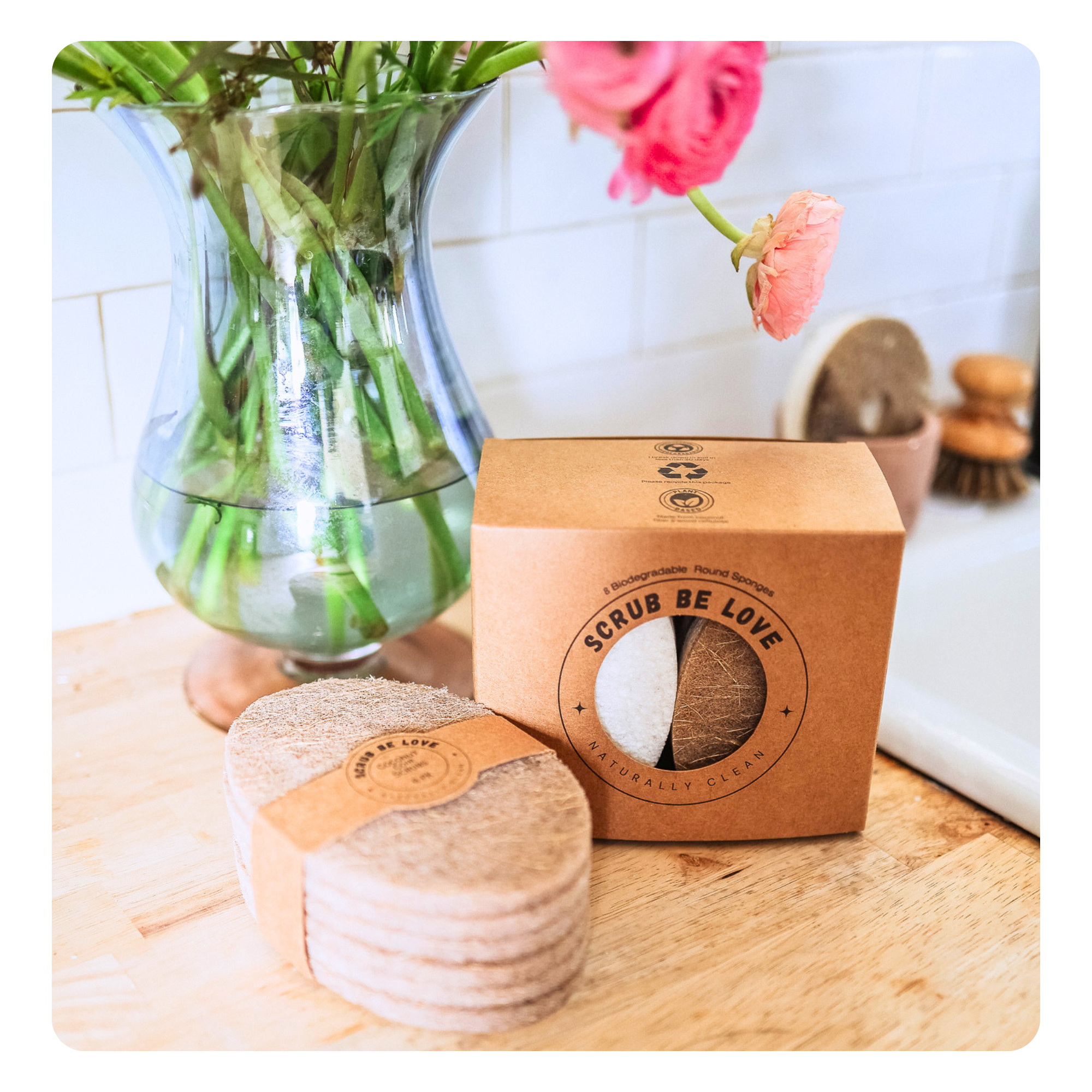A set of round natural fiber body scrubs in a kraft paper box labeled 'SCRUB BE LOVE', placed on a wooden surface next to a glass vase with pink flowers and green stems.