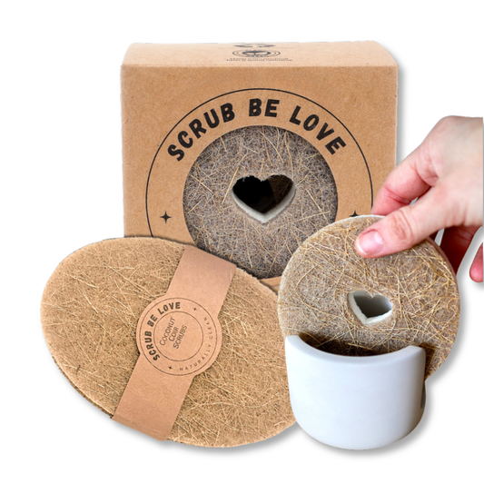 Two oval natural fiber scrubber pads with heart-shaped cutouts are shown, one resting on a white mug and one being held by a hand. A branded kraft box reading 'SCRUB BE LOVE' stands behind them on a white background.