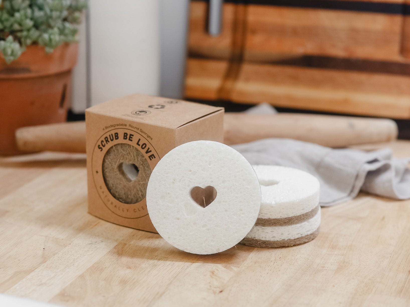 Natural plant-based biodegradable and compostable kitchen sponges sitting on top of a butcher block counter with a wooden rolling pin, linen dish towel, and wooden cutting board in the background.